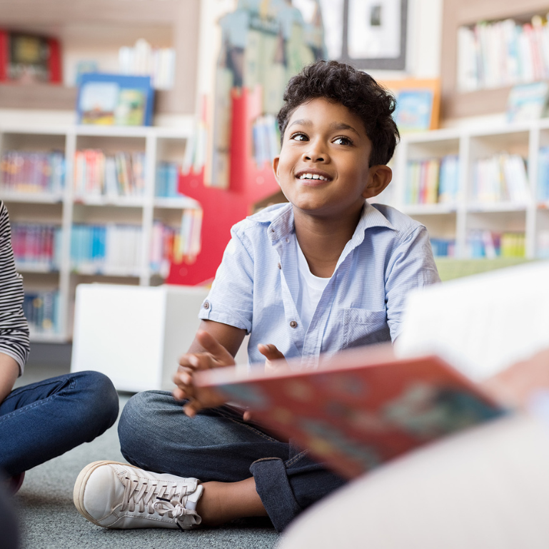 Smiling young boy sitting in a library reading area, engaged and attentive, with bookshelves in the background, promoting literacy and education for children.