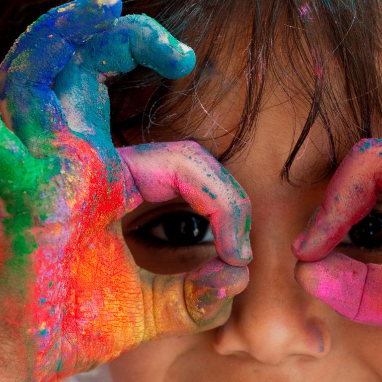Child with colorful paint-covered hands playfully framing their eyes, highlighting creativity and artistic expression in children