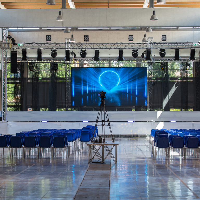 A large empty conference room with rows of blue chairs and a stage with a large screen