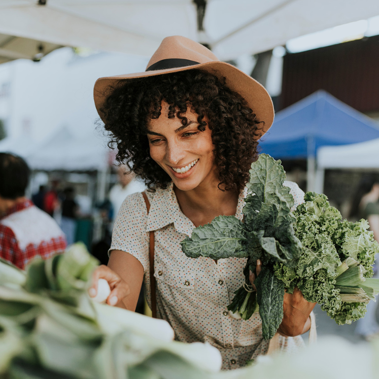 Woman with curly hair wearing a hat, smiling while selecting fresh greens at an outdoor market